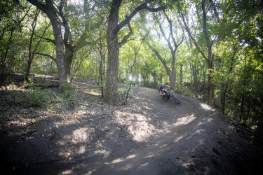 Mountain biker navigating a curved dirt trail surrounded by lush green trees and sunlight filtering through the foliage. Great Bear mountain bike trail.