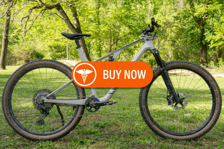 A modern mountain bike positioned on green grass, surrounded by trees. An orange "Buy Now" button with a medical symbol overlay is featured prominently.