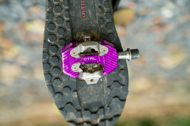A close-up view of a purple bicycle pedal attached to the sole of a cycling shoe, resting on a textured surface with visible dirt. The pedal features a silver mechanism and has "CTRL" inscribed on it. The shoe sole has a honeycomb pattern, indicating grip for cycling.