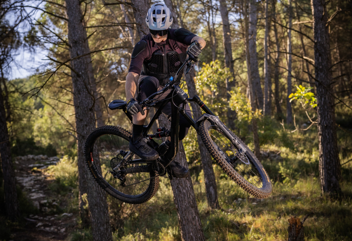 A cyclist performing a jump on a mountain bike, surrounded by trees in a forested area. The rider is wearing a helmet and protective gear, showcasing an action shot that captures the excitement of mountain biking. The scene is set in natural daylight, highlighting the greenery and rugged terrain.