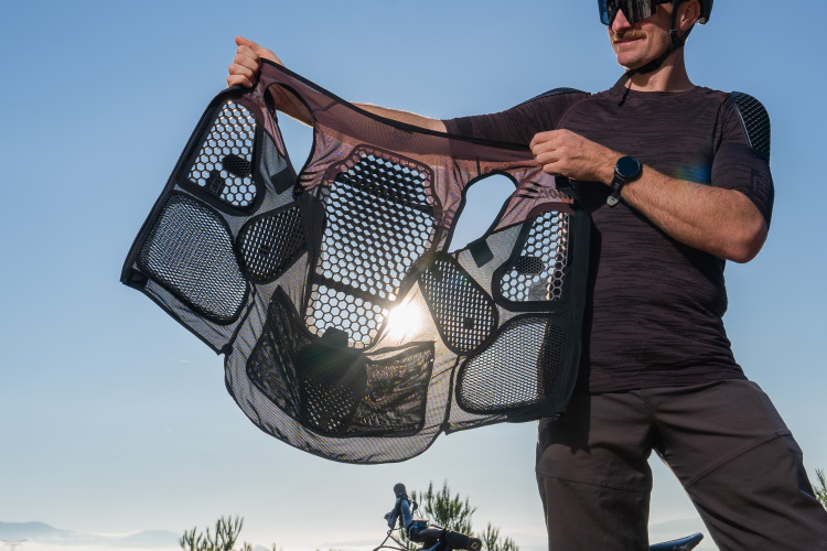 A person wearing a helmet and sunglasses holds up a lightweight, mesh bike protector against a clear blue sky, showcasing its unique design with honeycomb patterns. In the background, a bicycle and greenery are visible.