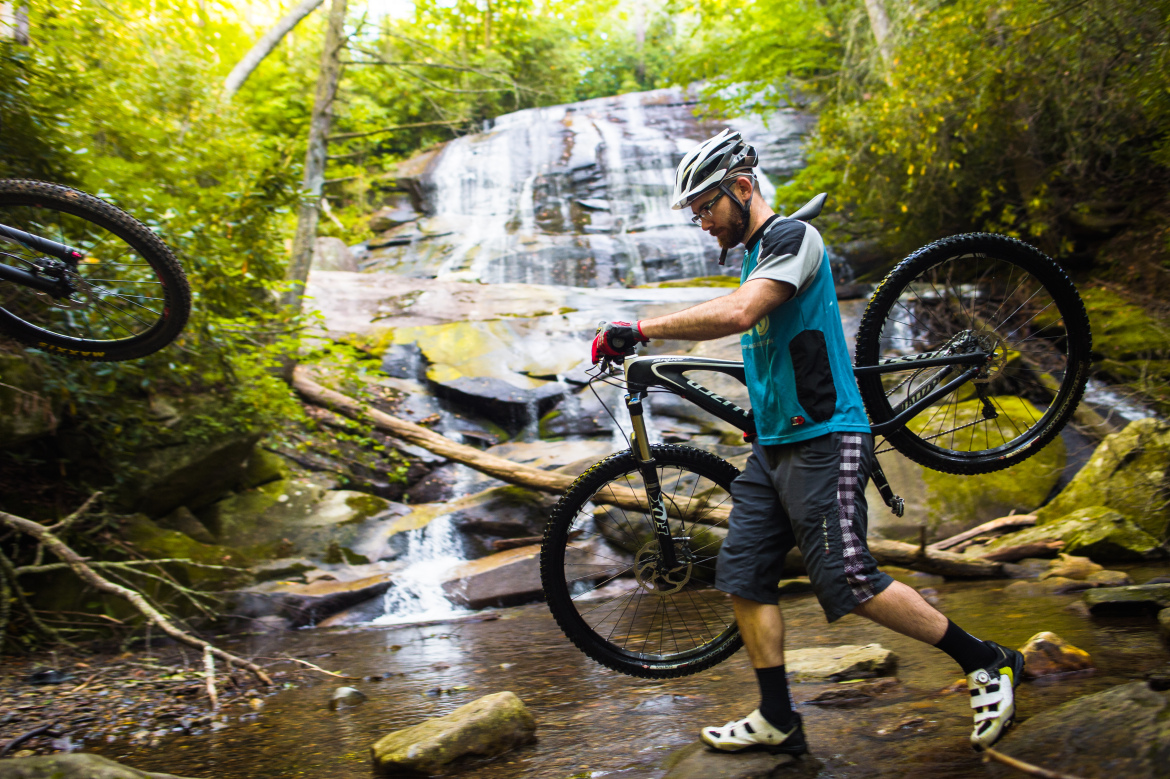 A cyclist carrying a mountain bike across a rocky stream, with a waterfall and lush greenery in the background. The cyclist is wearing a helmet and cycling gear, focusing on navigating the terrain.