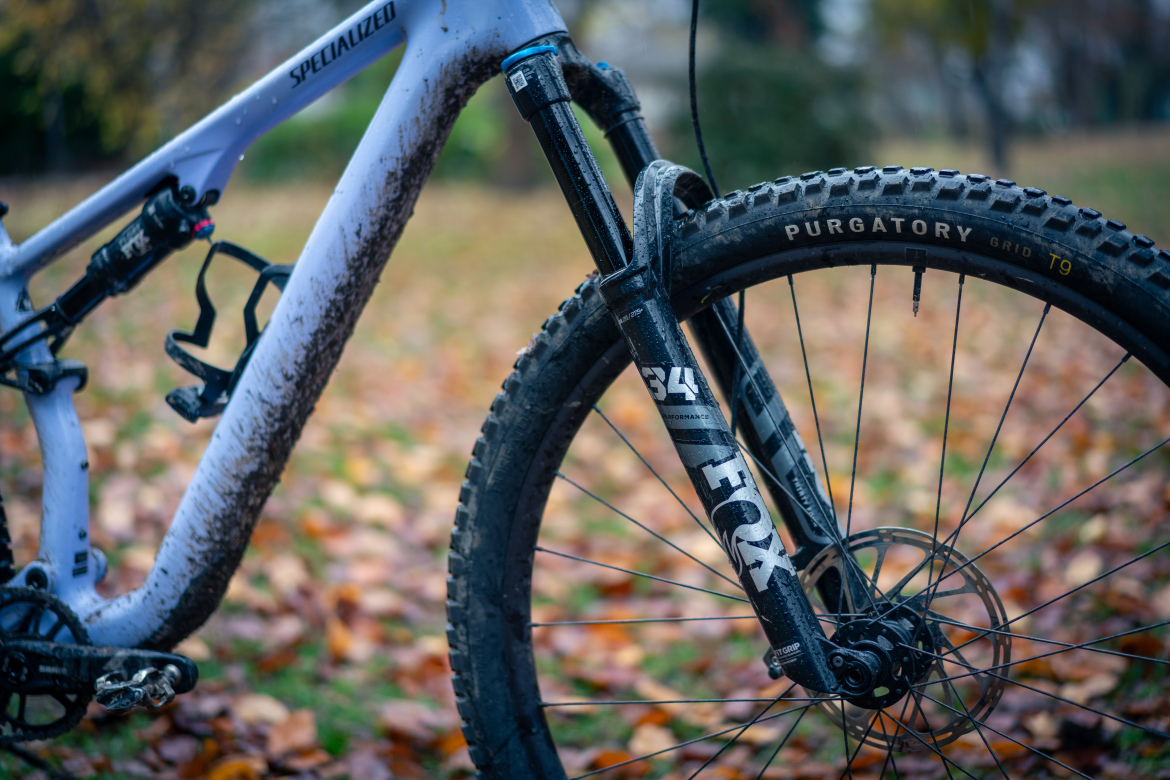 A close-up view of a mountain bike's front fork and tire, showcasing a specialized frame and muddy components. The tire features a tread pattern labeled "Purgatory," and the front fork is branded with "FOX" and model number "34." The background includes a carpet of fallen leaves, indicating an autumn setting.