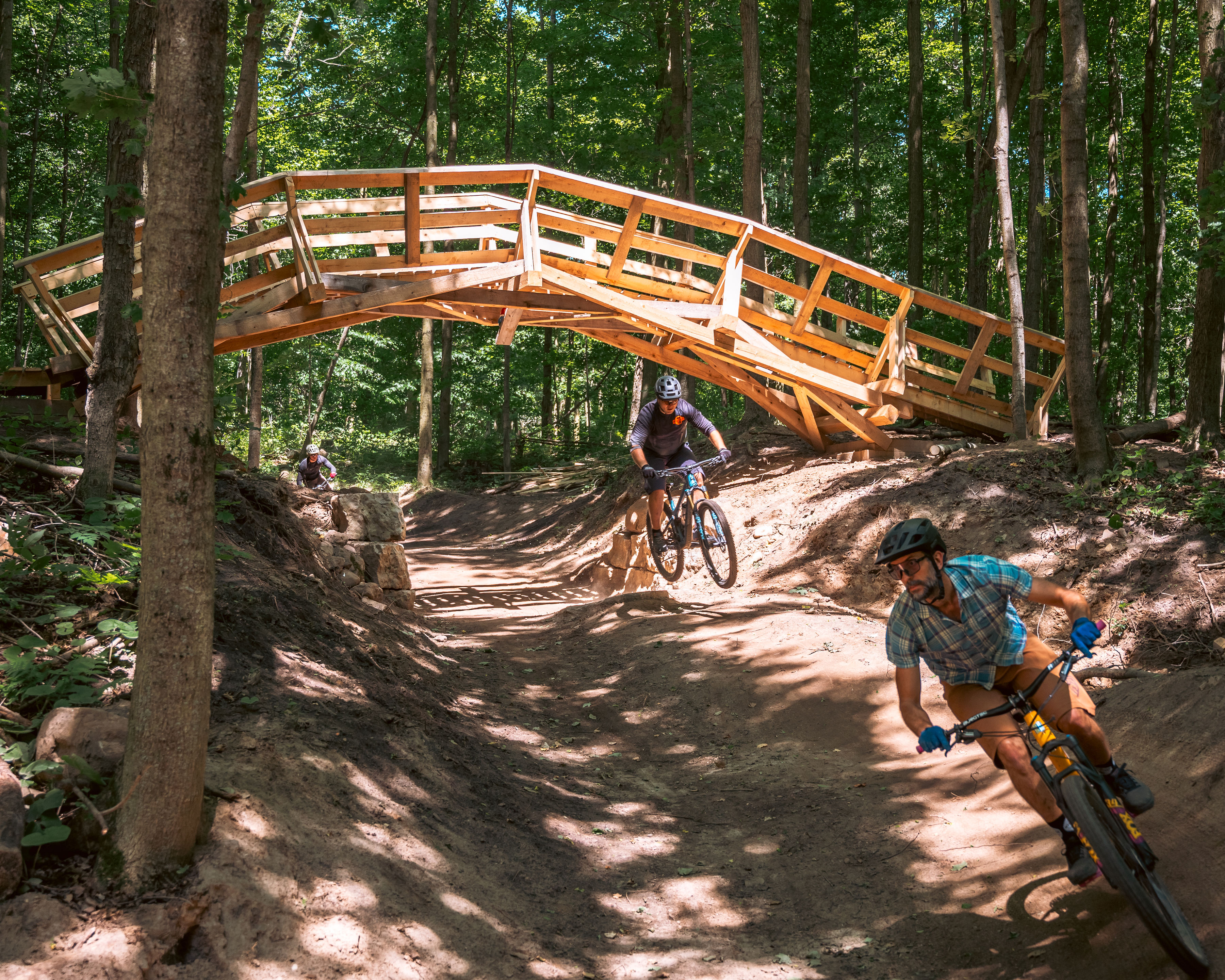 Two mountain bikers navigate a dirt trail through a wooded area, one riding closely beside a wooden bridge overhead. The scene is vibrant with greenery, and sunlight filters through the trees, highlighting the riders and the path. Hydrocut mountain bike trail.