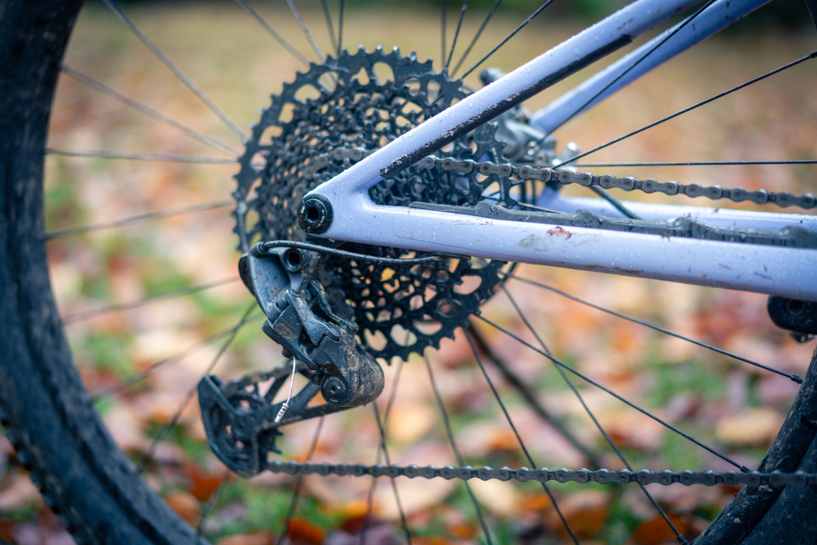 Close-up view of the rear derailleur and cassette of a mountain bike, with a muddy chain and a purple frame. The background features blurred autumn leaves on the ground.