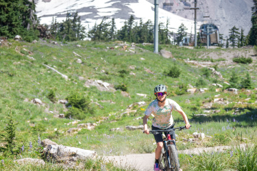 A mountain biker rides along a dirt trail in a scenic landscape, with lush green grass and wildflowers surrounding the path. In the background, a snow-capped mountain peak rises against a clear blue sky, and a ski lift is visible on the mountainside. Timberline Bike Park mountain bike trail.