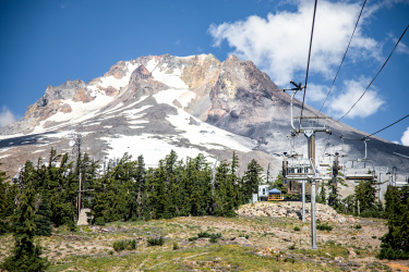 A scenic view of a mountain with patches of snow, surrounded by evergreen trees. In the foreground, a ski lift is visible, with several empty chairs and support poles. The sky is bright blue with a few clouds, enhancing the picturesque landscape. Timberline Bike Park mountain bike trail.