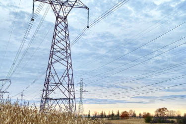 A tall, metal power transmission tower stands beside a dirt path, surrounded by fields with dried corn stalks. The sky above is cloudy, with hints of orange and blue during sunset, while additional power lines stretch across the horizon. Lafortune Park mountain bike trail.