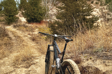 A black mountain bike parked on a sandy trail surrounded by tall grass and sparse trees under a cloudy sky. Lambton County Heritage Forest mountain bike trail.