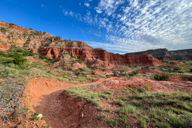 A scenic view of a rugged landscape featuring red rock formations and green vegetation under a blue sky with scattered clouds. A winding dirt path leads through the foreground, inviting exploration of the natural formations in the background.