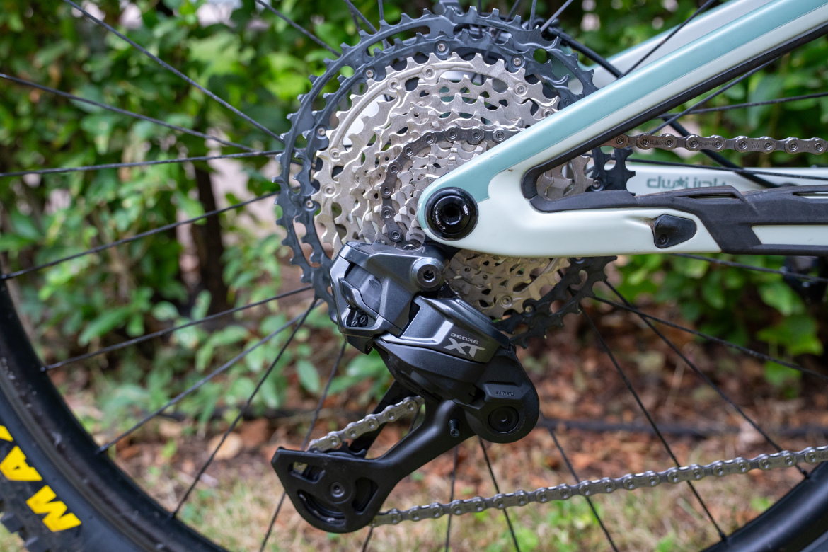 Close-up of a bicycle's rear drivetrain, featuring a cluster of gears and a derailleur. The derailleur is marked with "Deore XT" and is mounted on a light-colored bicycle frame. The background includes greenery, suggesting an outdoor setting.