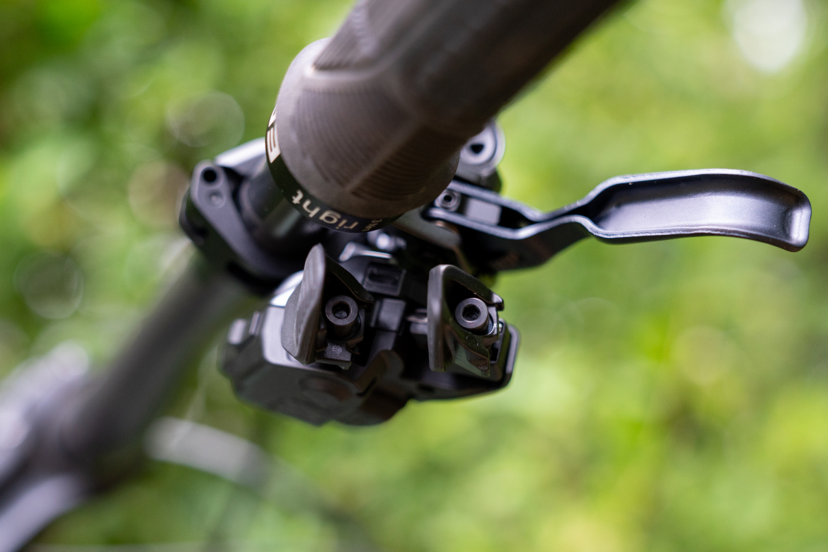 Close-up of a bicycle handlebar component featuring a shifter or lever mechanism, with a textured grip and a blurred green background of foliage.