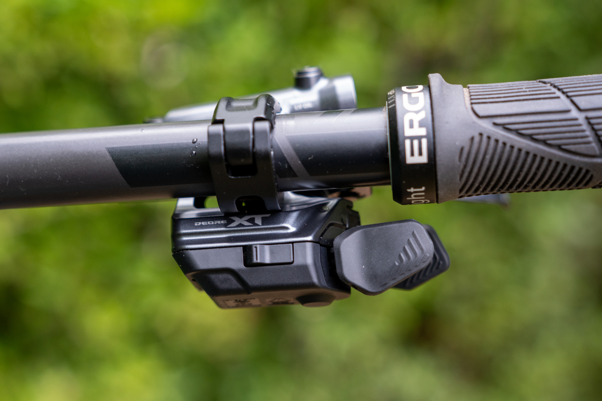 Close-up view of a bicycle handlebar featuring a Shimano Deore XT shifter and ergonomic grip, set against a blurred green background. The image highlights the sleek design of the shifter and grip, showcasing details such as texture and fastening mechanism.