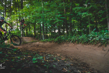Two mountain bikers performing tricks on a dirt track in a wooded area. One rider is jumping off a small ramp while the other is riding along a curved path, surrounded by lush green trees and foliage. Hydrocut mountain bike trail.