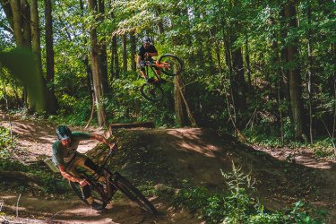 Two mountain bikers navigating a dirt bike trail in a lush green forest. One rider is making a sharp turn on the left, while the other is performing a jump above a dirt mound in the background, surrounded by trees and foliage. Sunlight filters through the leaves, highlighting the dynamic movement and natural setting. Hydrocut mountain bike trail.