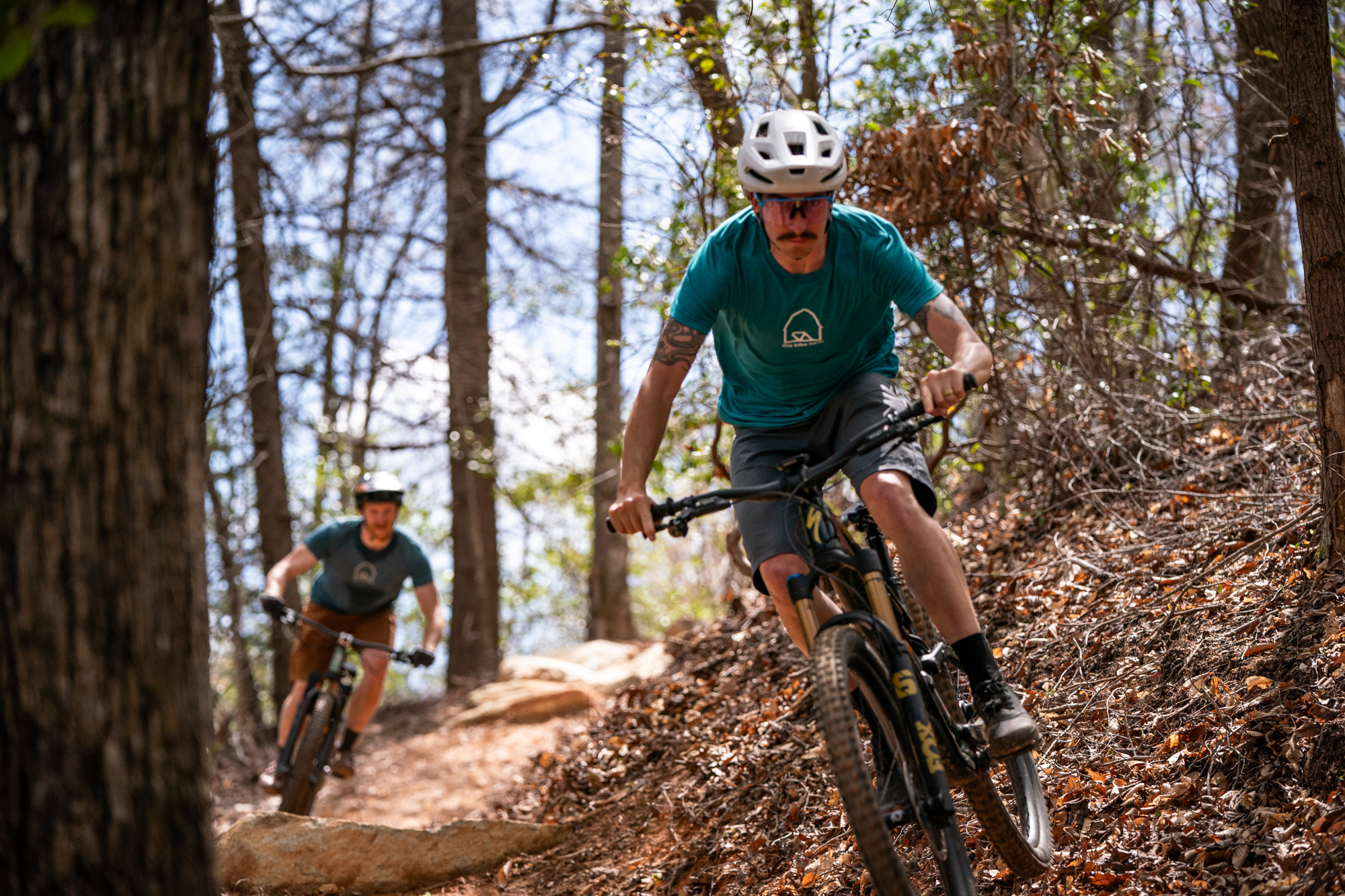 Two mountain bikers riding along a winding dirt trail through a wooded area. The foreground biker is leaning forward, wearing a teal shirt and a white helmet, while the background biker follows closely. Sunlight filters through the trees, illuminating patches of the path covered with fallen leaves and rocks. Foothills Watershed mountain bike trail.