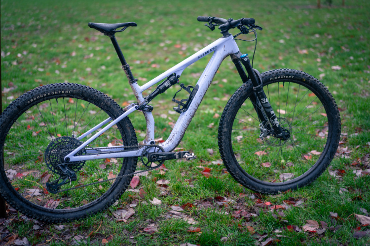 A mountain bike with a white frame and black components, resting on grass covered in fallen leaves. The bike is slightly muddy, indicating recent use on rugged terrain. The tires display a pronounced tread pattern suitable for off-road riding.