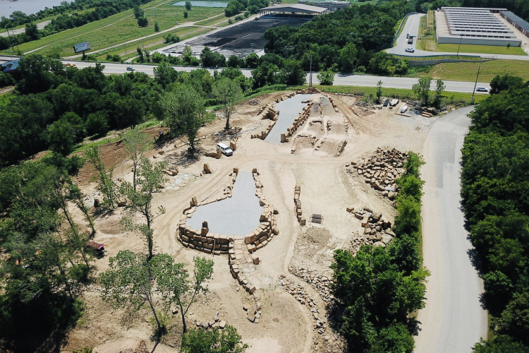 Aerial view of a construction site surrounded by trees, featuring a cleared area with stone piles and gravel. In the background, a roadway runs alongside the site, with additional structures and greenery visible, indicating an urban or suburban setting.