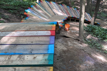 A colorful wooden ramp spirals through a forested area, made up of varying shades of paint on the planks. The ramp is situated on a dirt path surrounded by trees and greenery. A Home Depot bucket is visible nearby, suggesting ongoing construction or maintenance. Hydrocut mountain bike trail.