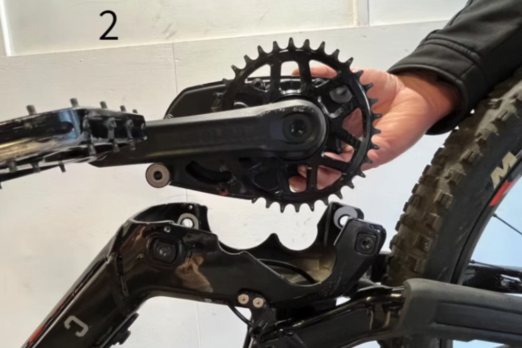 A close-up image of a bicycle's chainring and crankset, showing a hand gripping the crank arm. The bicycle frame is black, and the background features a plain white wall. A label in the top left corner indicates the step number as "2."