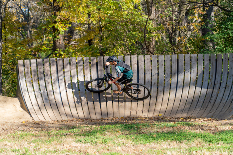 A young person riding a mountain bike on a wooden ramp in a park, surrounded by trees with autumn-colored leaves. The rider is wearing a helmet and is positioned mid-action, leaning into the curve of the ramp.