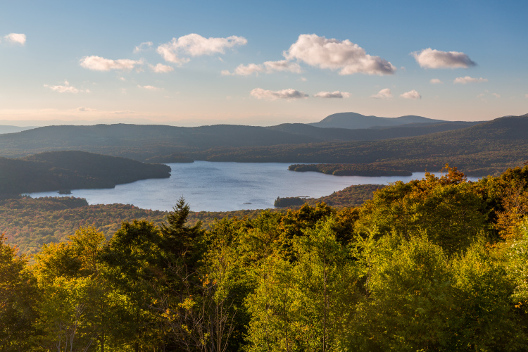 A panoramic view of a serene lake surrounded by rolling hills and vibrant autumn foliage under a clear blue sky with a few clouds. The scene captures the beauty of nature in a tranquil setting.