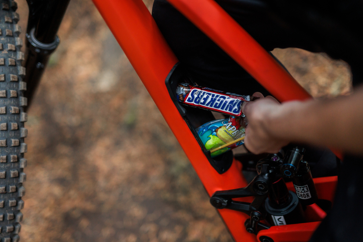 A hand reaching into the frame of a bright orange mountain bike, retrieving snacks including a Snickers bar and a Capri Sun drink pouch. The background features blurred natural scenery, suggesting an outdoor setting.