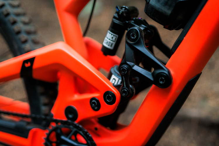 Close-up view of a brightly colored orange mountain bike frame, showcasing the intricate suspension system and components, including black linkage and shock absorber details, with a blurred background.