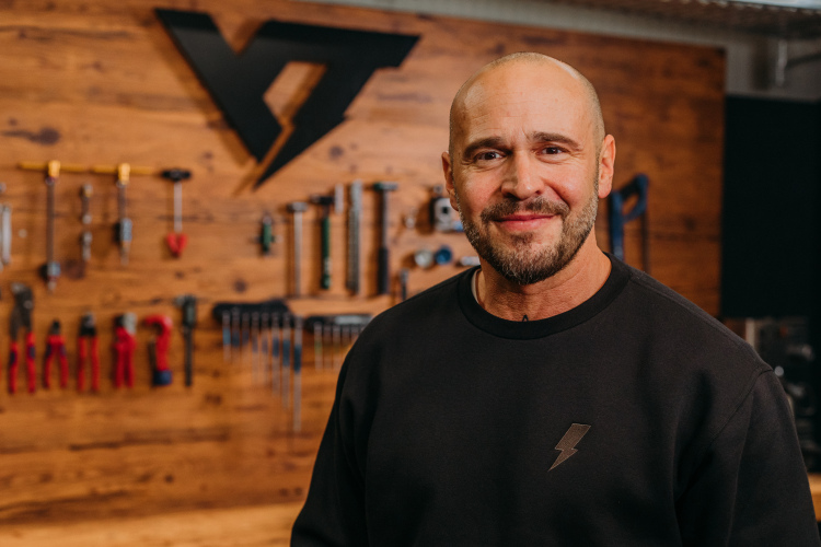 A smiling man with a beard is standing in front of a wooden wall adorned with various tools and a logo. He is wearing a black sweatshirt featuring a lightning bolt design. The atmosphere suggests a workshop or studio setting.