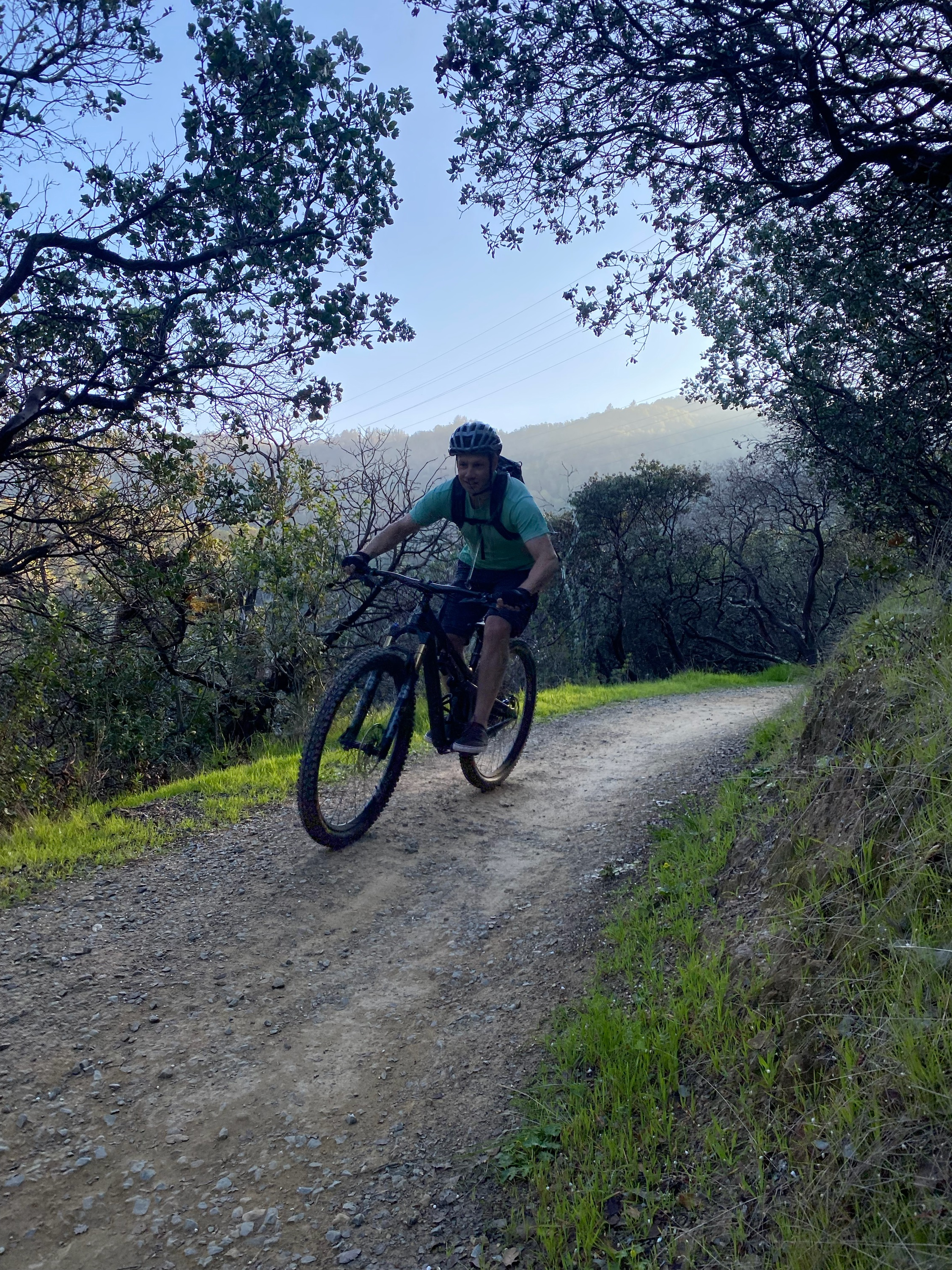 A person riding a mountain bike on a gravel trail surrounded by trees and greenery, with a slight incline leading into the background. The scene is illuminated by soft morning light, creating a serene outdoor atmosphere.
