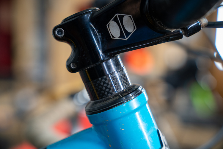 Close-up of a bike's headset showing detailed features, including a black stem with a logo, a carbon fiber section, and a blue frame beneath. The background is slightly blurred, suggesting a workshop or garage environment filled with bike tools and parts.