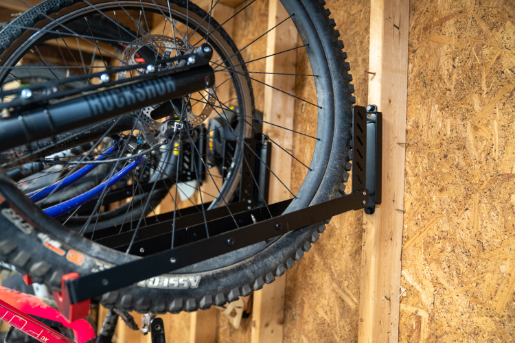 A close-up view of a mountain bike wheel mounted on a black wall rack against an oriented strand board wall. The rack securely holds the wheel, showcasing the tire and disc brake. Other colorful bike components are partially visible in the background.