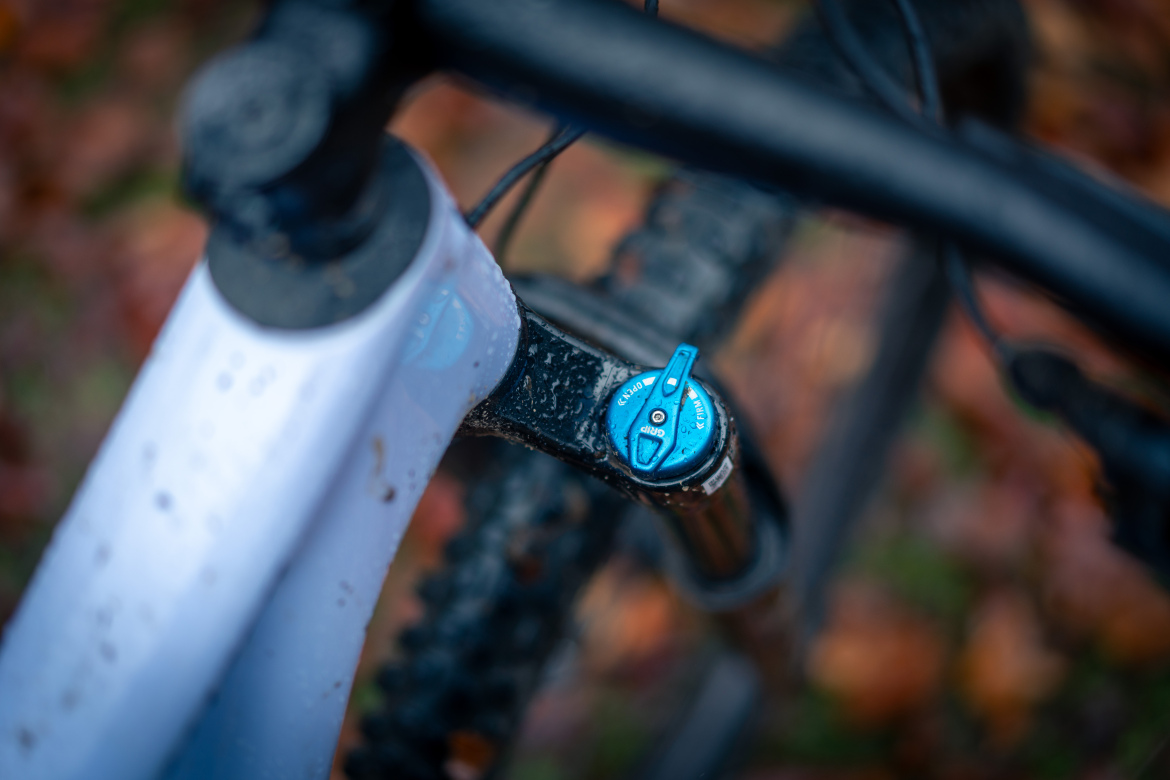 Close-up view of a mountain bike's suspension system, featuring a blue adjustment knob on a black fork, with raindrops on the surface, surrounded by blurred autumn leaves in the background.
