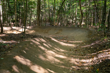 A dirt bike trail winding through a shaded forest, featuring a series of smooth, raised berms and gentle curves, surrounded by green trees and scattered autumn leaves. Kingdom Trails mountain bike trail.