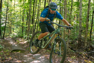 A mountain biker performing a jump on a dirt trail surrounded by lush green trees and sunlight filtering through the canopy. The cyclist is wearing a helmet, sunglasses, and a blue shirt, with one wheel off the ground as they navigate the terrain. Kingdom Trails mountain bike trail.