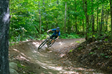 A mountain biker navigates a curved dirt trail through a lush, green forest, demonstrating speed and agility. Sunlight filters through the leaves above, highlighting the vibrant surroundings. Kingdom Trails mountain bike trail.