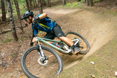 A cyclist in a blue jersey and black shorts leans into a sharp turn while riding a mountain bike on a dirt trail surrounded by tall trees. The forest is filled with green foliage and scattered leaves on the ground, creating a natural biking environment. Kingdom Trails mountain bike trail.