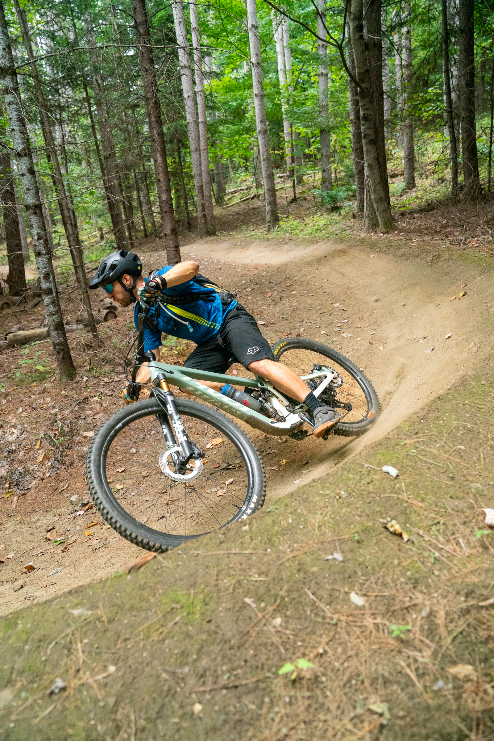A cyclist in a blue jersey and black shorts leans into a sharp turn while riding a mountain bike on a dirt trail surrounded by tall trees. The forest is filled with green foliage and scattered leaves on the ground, creating a natural biking environment. Kingdom Trails mountain bike trail.