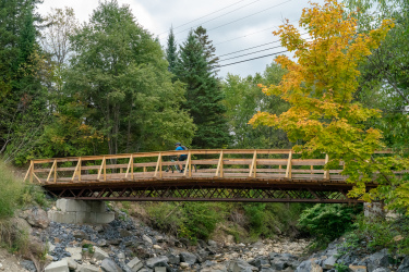 A wooden bridge spans a rocky creek surrounded by vibrant trees, including some with autumn-colored leaves. A cyclist in blue rides across the bridge, set against a backdrop of green foliage and a cloudy sky. Kingdom Trails mountain bike trail.