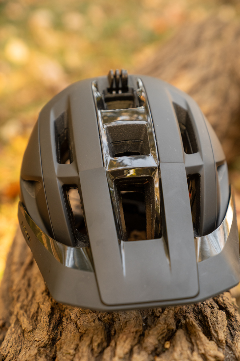 A close-up view of a black bicycle helmet placed on a wooden surface, with autumn foliage blurred in the background. The helmet features multiple ventilation openings on the top, showcasing its aerodynamic design.