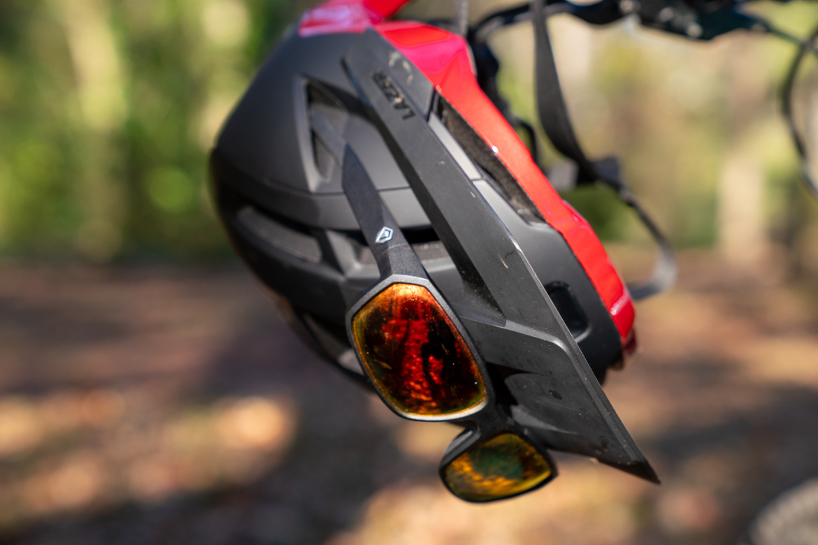 A close-up of a brightly colored bicycle helmet hanging with a pair of sunglasses attached to it. The helmet features a black and red design, showcasing vents and straps. The background is a blurred natural setting with greenery.