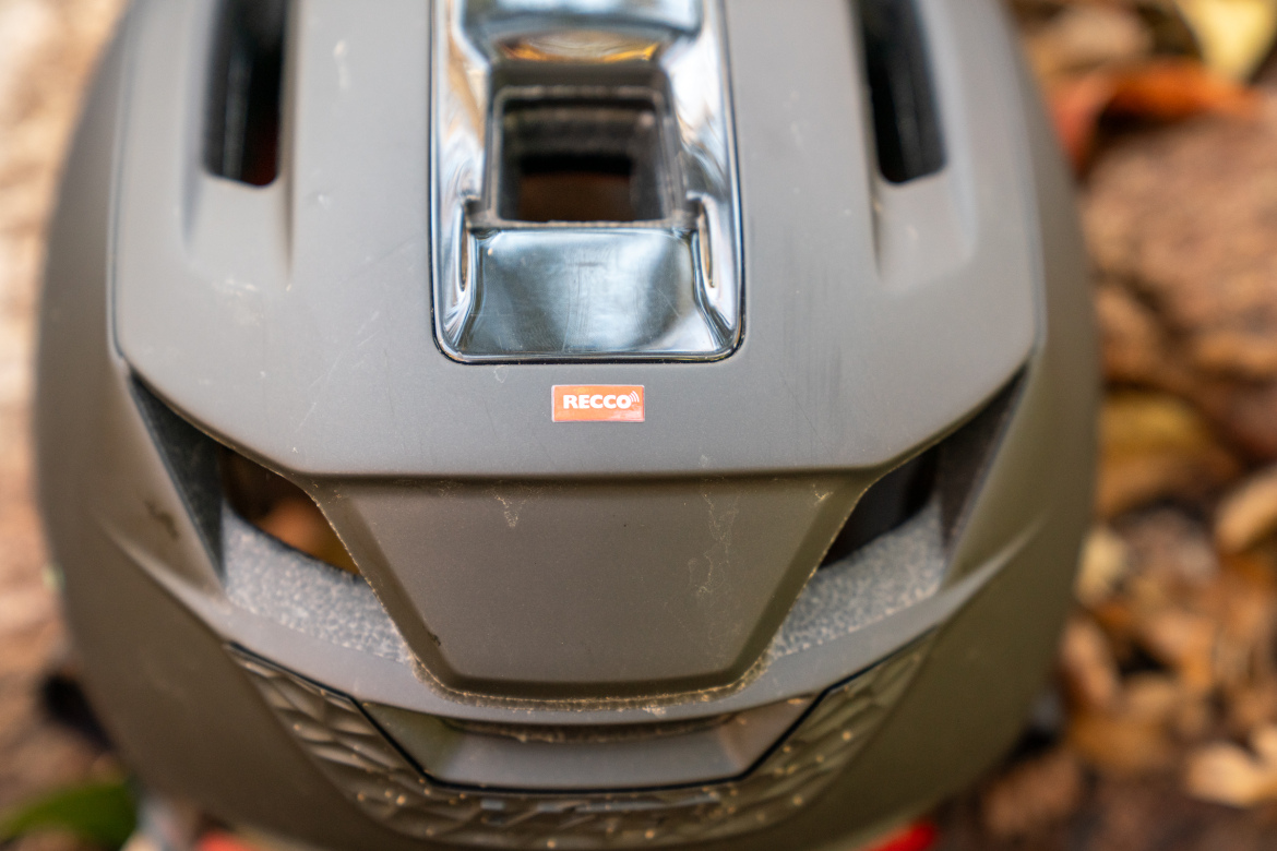 A close-up image of the top of a black helmet featuring a prominent RECCO® safety label. The helmet shows signs of use, with some dust and scratches on its surface. The background consists of blurred natural elements, suggesting an outdoor environment.