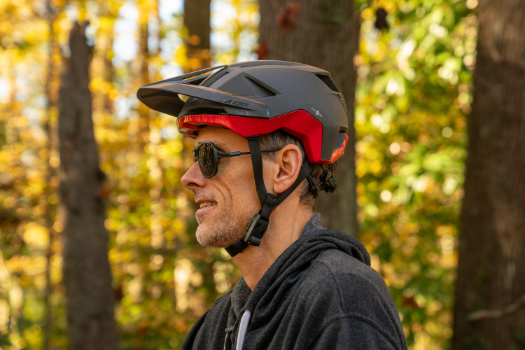A man wearing a black and red bike helmet and sunglasses stands outdoors with a blurred background of colorful autumn foliage and trees. He has short hair and a stubbly beard, and he appears to be looking off to the side. The image captures a moment of contemplation in nature.