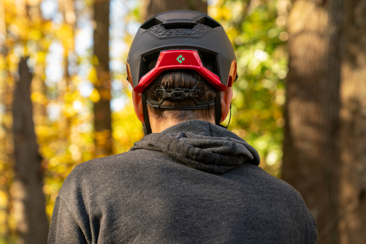 A person wearing a black and red helmet stands in a forest, seen from behind. They are dressed in a dark hoodie, and the background features blurred trees with autumn foliage.