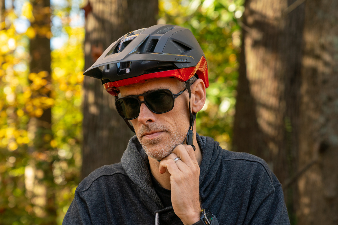 A person wearing a black and red bicycle helmet and sunglasses, touching their chin thoughtfully. The background features trees with autumn foliage, creating a vibrant outdoor scene. The individual is dressed in a dark hoodie, and their expression appears focused.