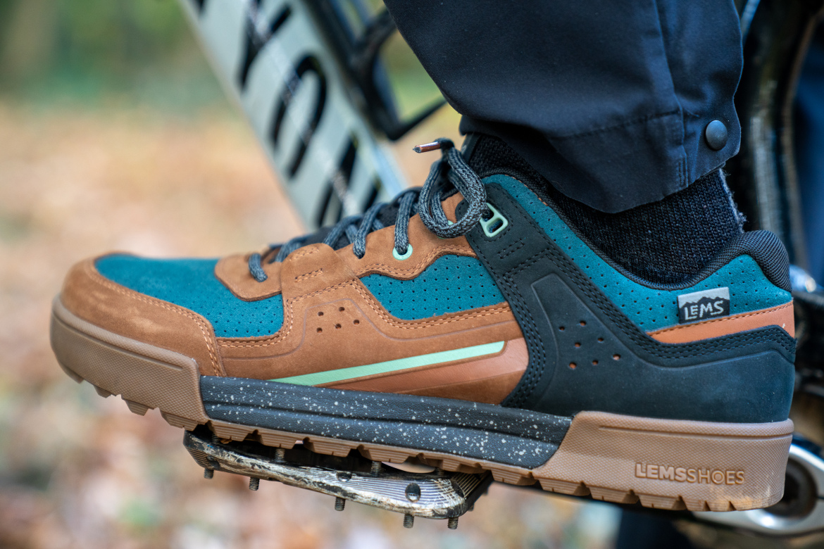 A close-up of a person's foot wearing a brown and teal sneaker while resting on a bicycle pedal. The sneaker features a mix of materials, including suede and mesh, with a speckled sole and branding visible on the side. The background shows a blurred natural setting with autumn leaves.