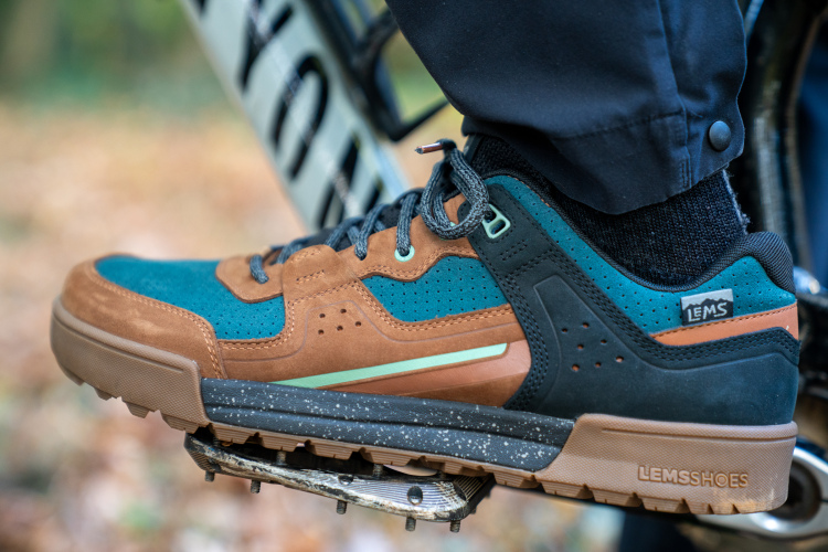 A close-up of a person's foot wearing a brown and teal sneaker while resting on a bicycle pedal. The sneaker features a mix of materials, including suede and mesh, with a speckled sole and branding visible on the side. The background shows a blurred natural setting with autumn leaves.