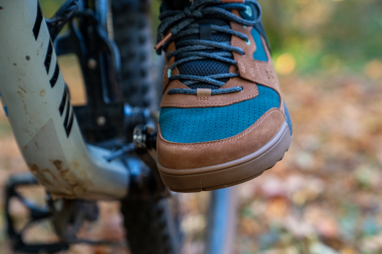 A close-up of a brown and teal sneaker resting on the pedal of a mountain bike, with a blurred background of autumn foliage. The shoe features a textured design and laces, highlighting an active outdoor setting.