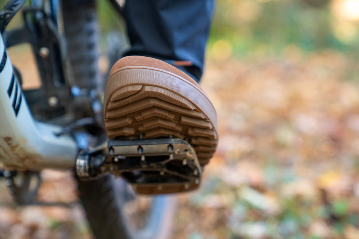 Close-up of a rubber-soled shoe resting on a bicycle pedal, with a blurred background of autumn leaves. The shoe has a distinctive tread pattern, highlighting its grip and traction.