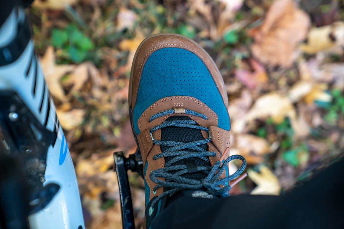 A close-up view of a brown and blue athletic shoe resting on a bicycle pedal, with a background of colorful autumn leaves on the ground.